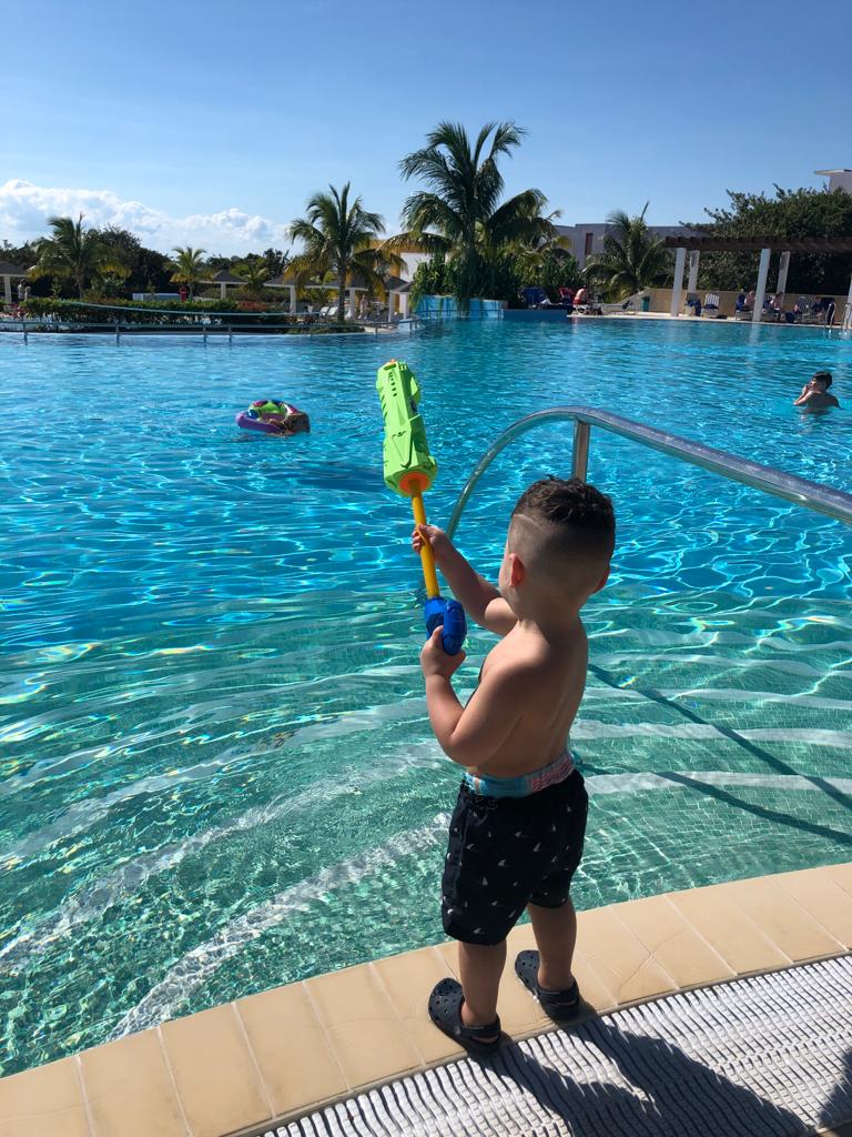boy toddler standing by the pool and spraying with a water gun. beautiful palm trees in the background